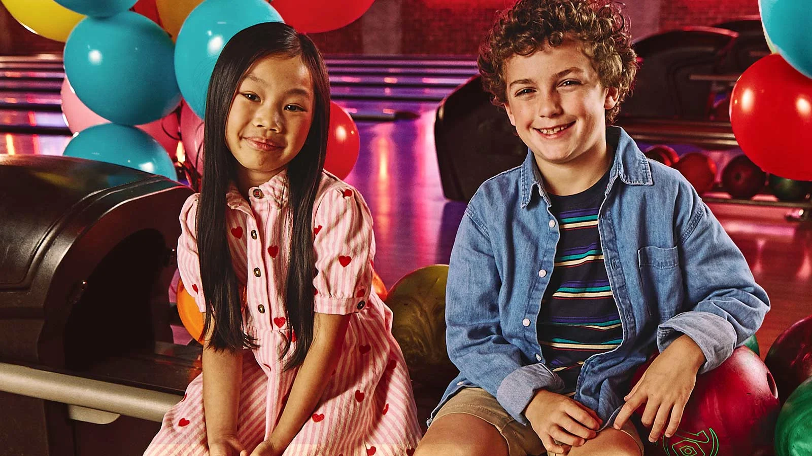 Two kids sitting in front of bowling lanes with balloons in the background