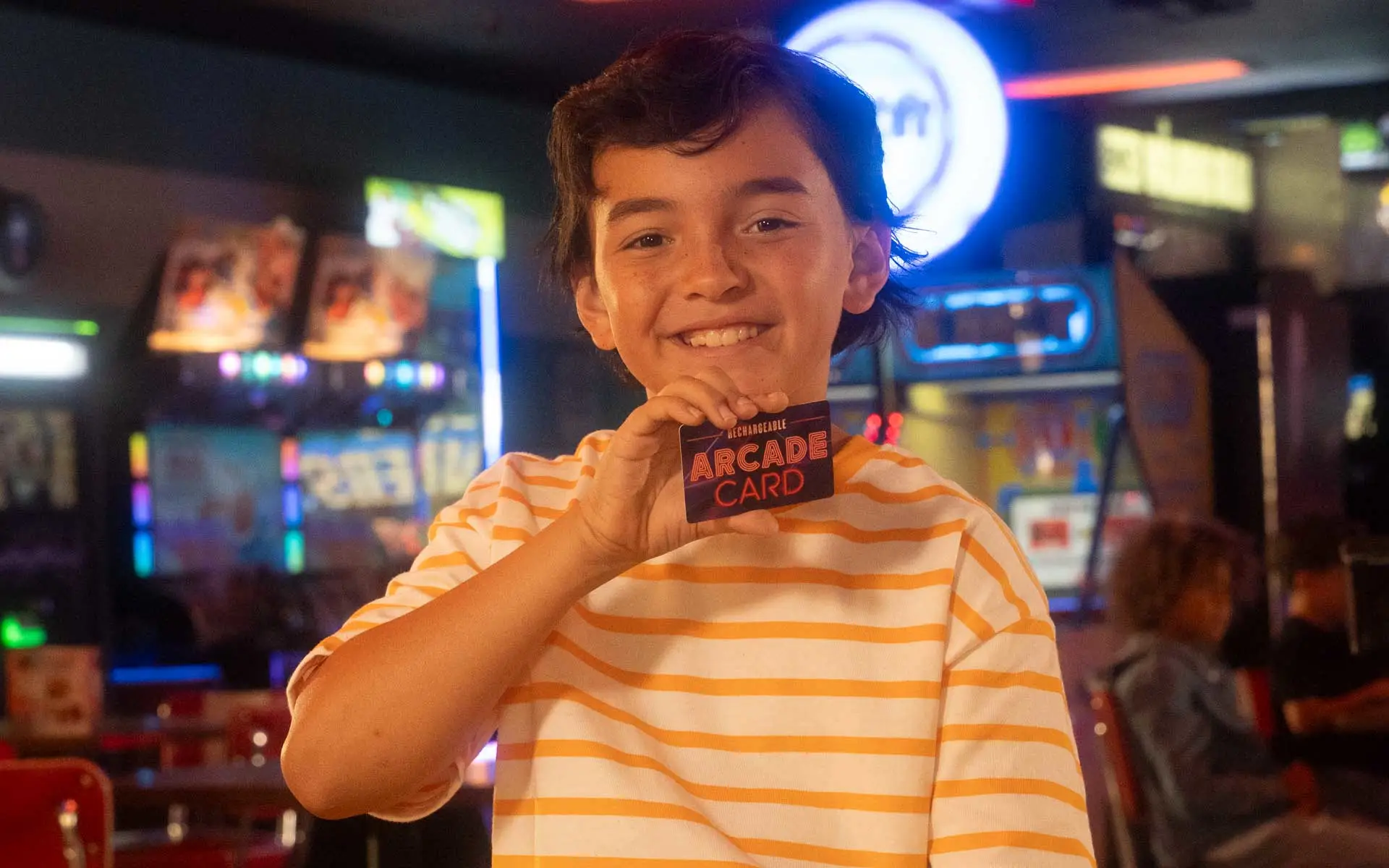 Smiling child holding an arcade game card inside a family entertainment center with colorful arcade machines and neon lights.
