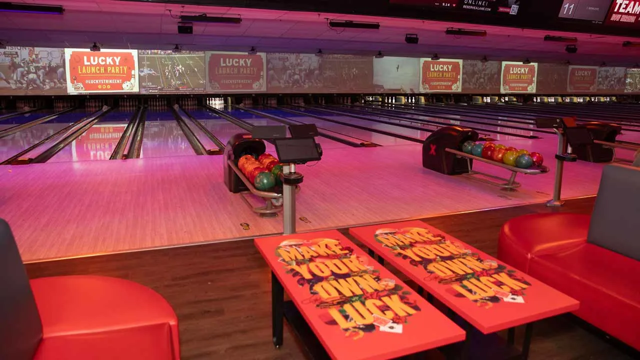Pink-lit bowling lanes at Lucky Strike Boca Raton with red lounge seating, 'Make Your Own Luck' tabletops, and Lucky Launch Party signage on video walls