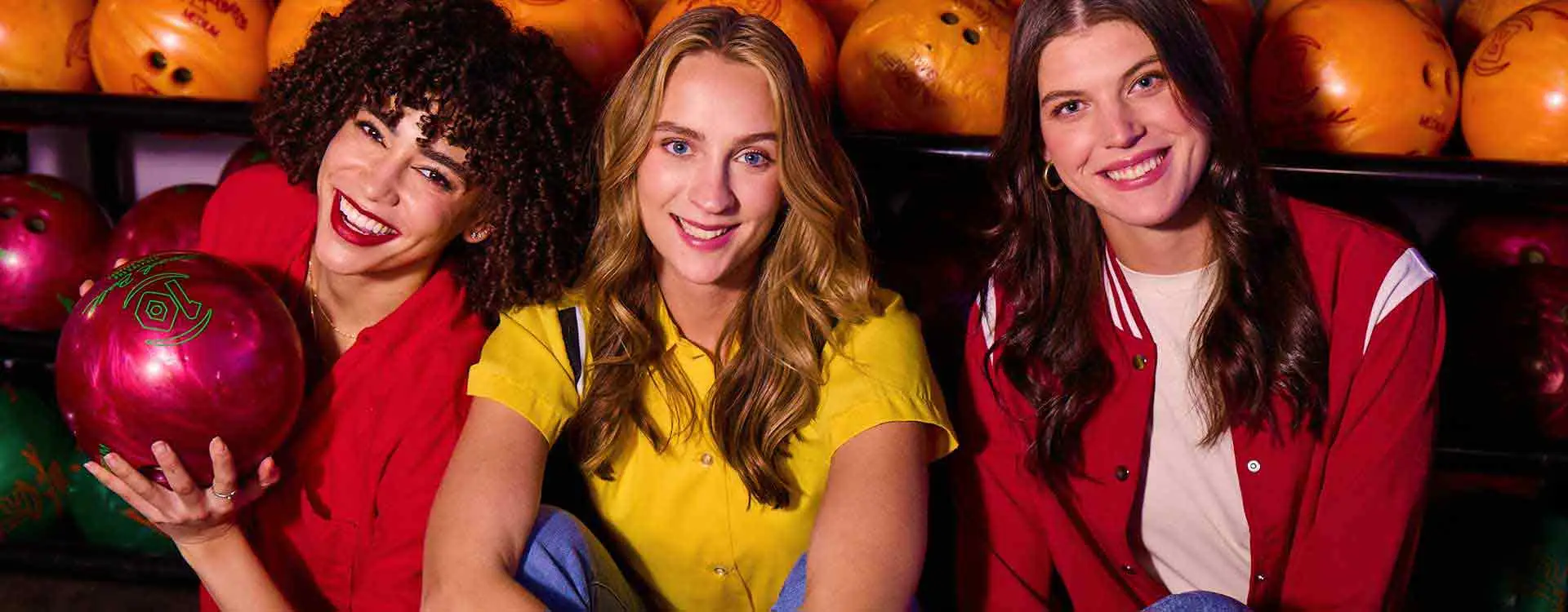 Three women smiling in front of a bowling ball rack at AMF Bowling, ready for a fun night out.