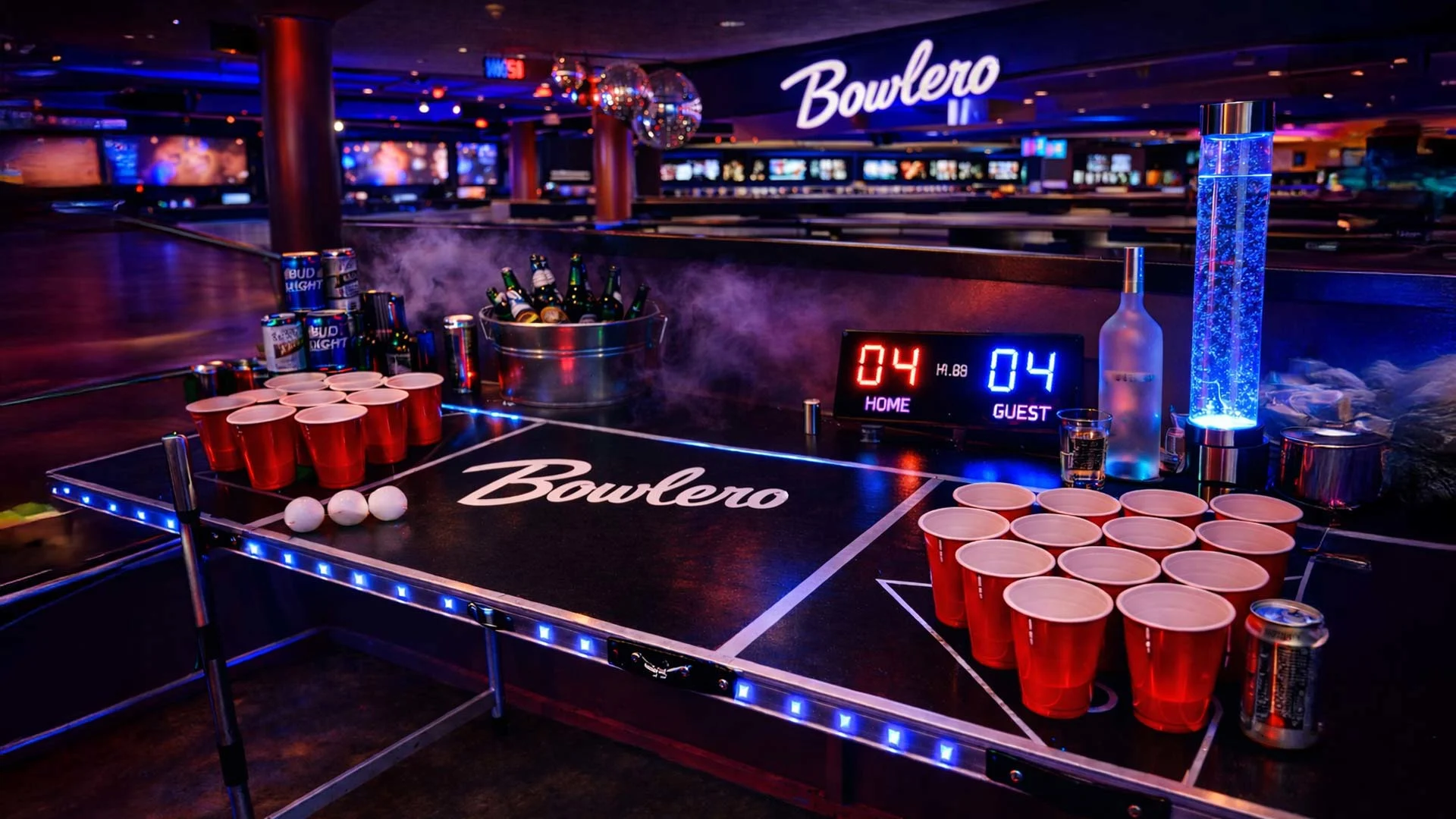 A neon-lit beer pong table set up inside a modern bowling entertainment venue, with red plastic cups arranged on both ends, ping-pong balls on the table, and bowling lanes and glowing signage visible in the background under colorful arcade-style lighting.