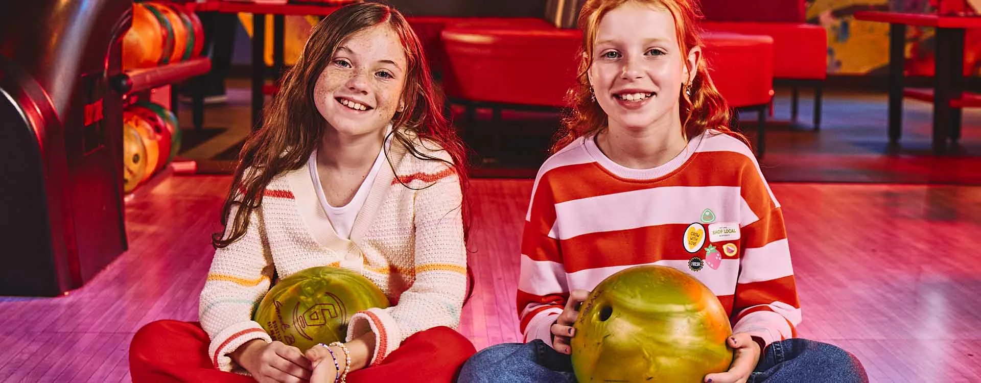 Two girls with bowling balls and a bowling alley in the background
