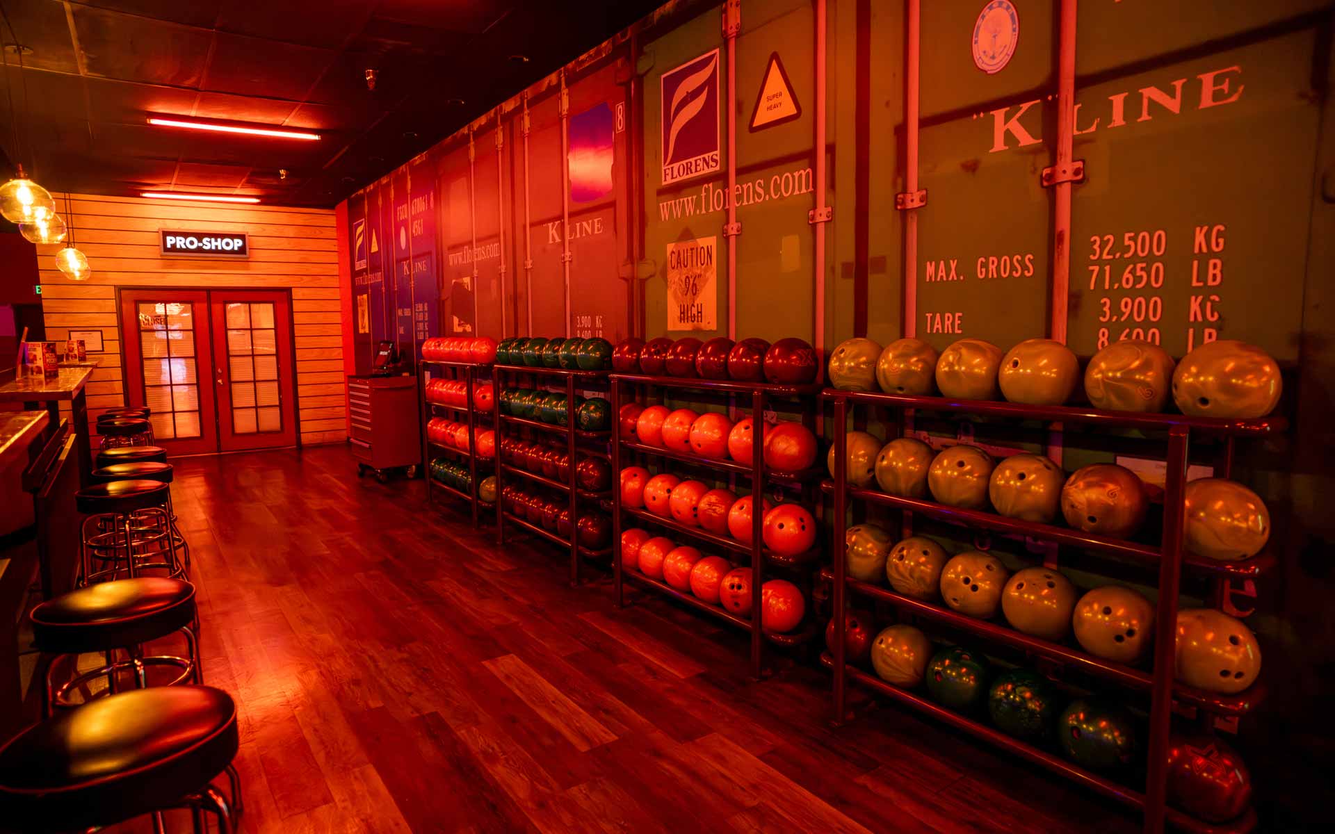 Bowling pro shop area with rows of colorful bowling balls on racks, industrial-style decor, and warm ambient lighting.