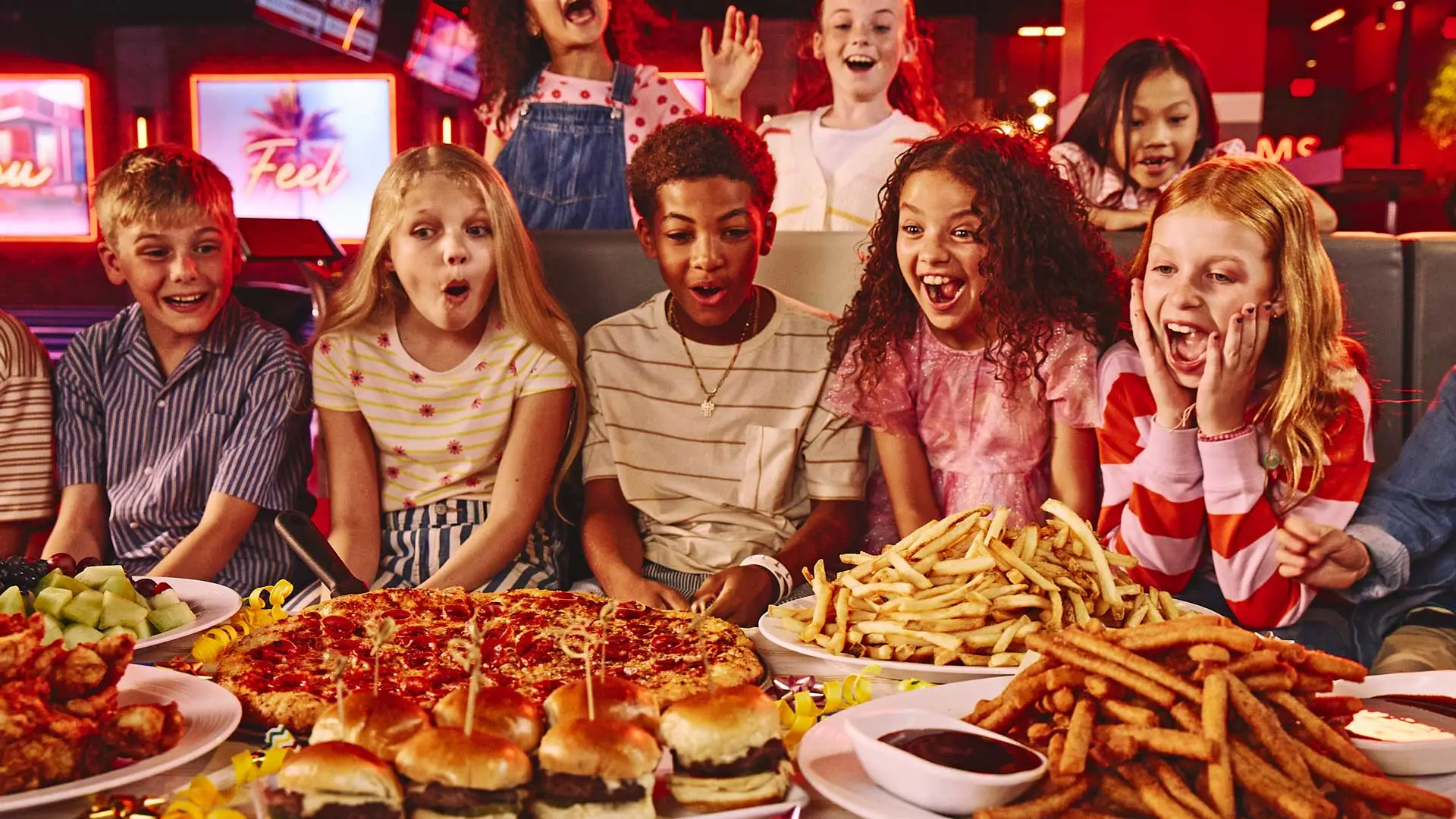 Kids excitedly gather around a table filled with party food—including pizza, fries, sliders, wings, and fruit—at a colorful bowling entertainment venue.