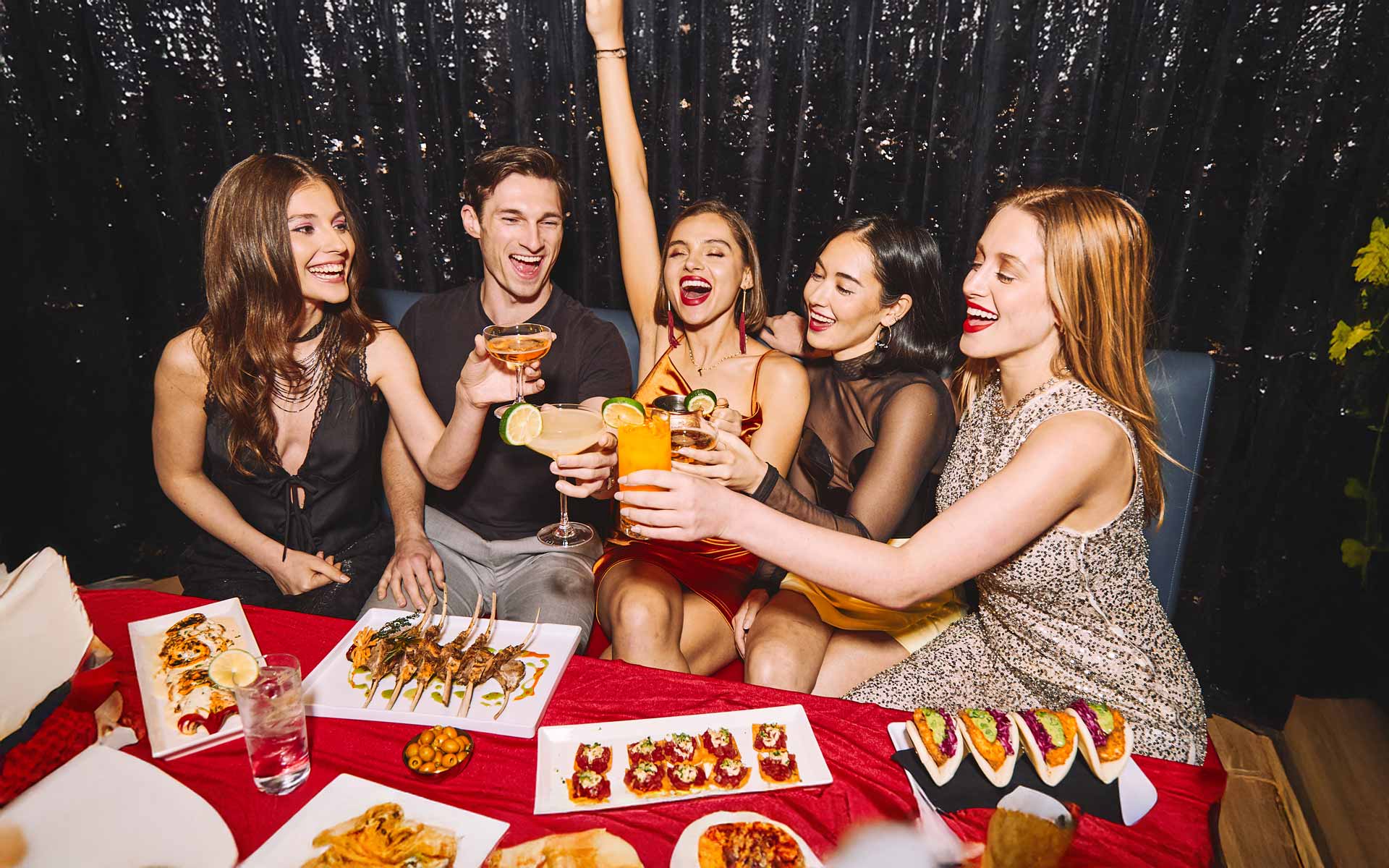 Group of friends dressed for a night out raising cocktails and celebrating together at a table with appetizers and drinks.