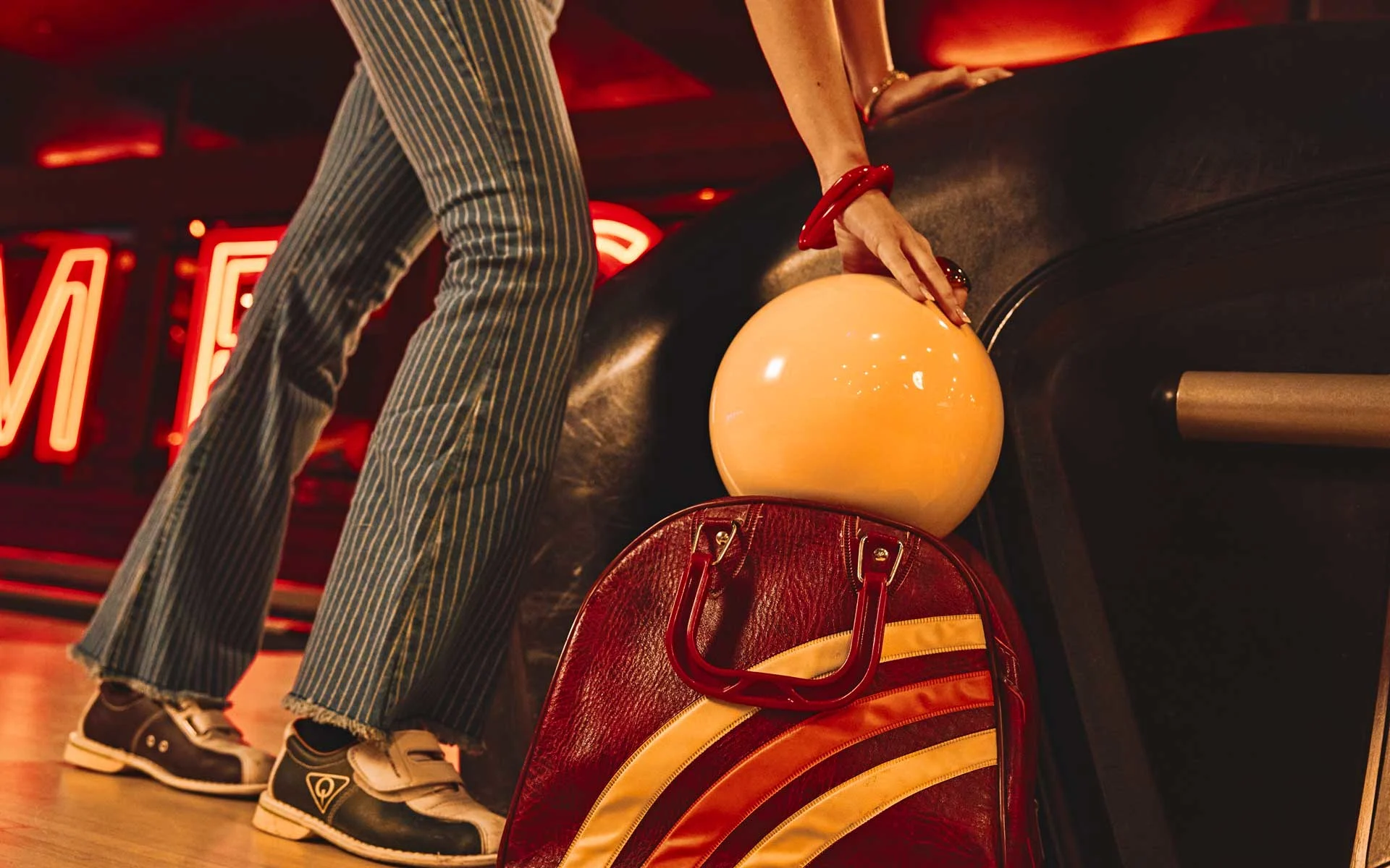 Retro-inspired bowling scene with a person placing a cream-colored bowling ball into a red leather bag, wearing striped pants and classic bowling shoes, set against warm neon lighting in a stylish, upscale bowling alley.