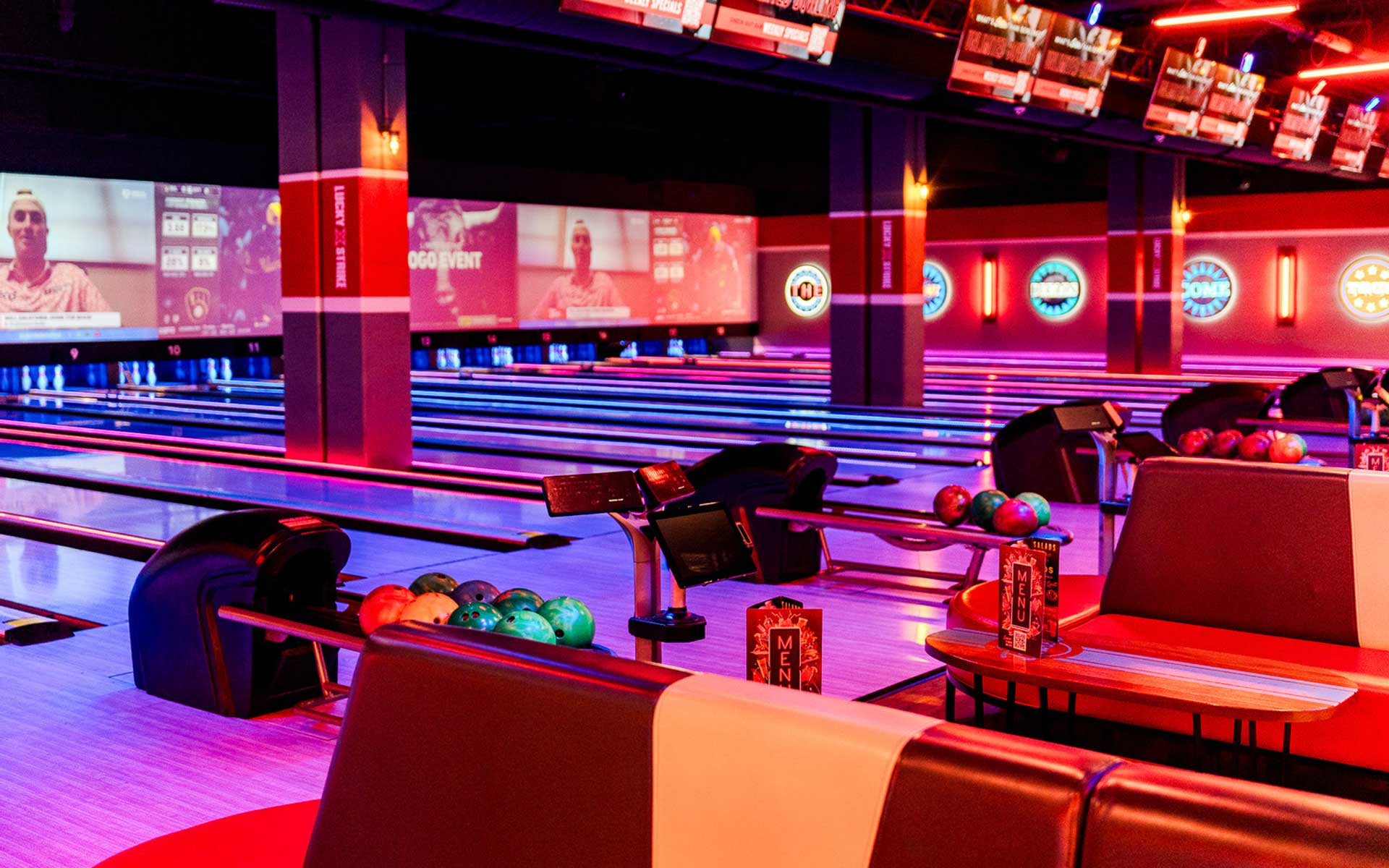 Neon-lit bowling alley featuring multiple lanes, colorful ball racks, and lounge-style seating, with large projection screens and vibrant red and blue lighting creating a high-energy, modern entertainment atmosphere.