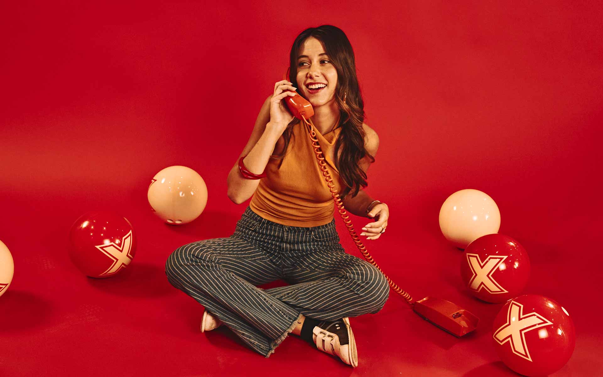 Woman sitting on a red backdrop talking on a retro phone, surrounded by bowling balls with “X” logos, creating a playful, stylish, and nostalgic scene.