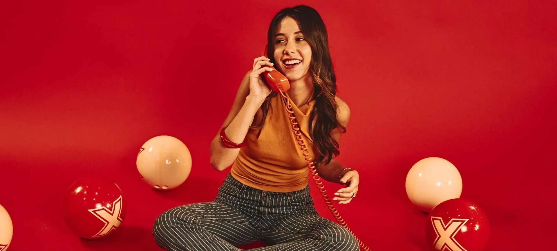 Woman sitting on a red backdrop talking on a retro phone, surrounded by bowling balls with “X” logos, creating a playful, stylish, and nostalgic scene.