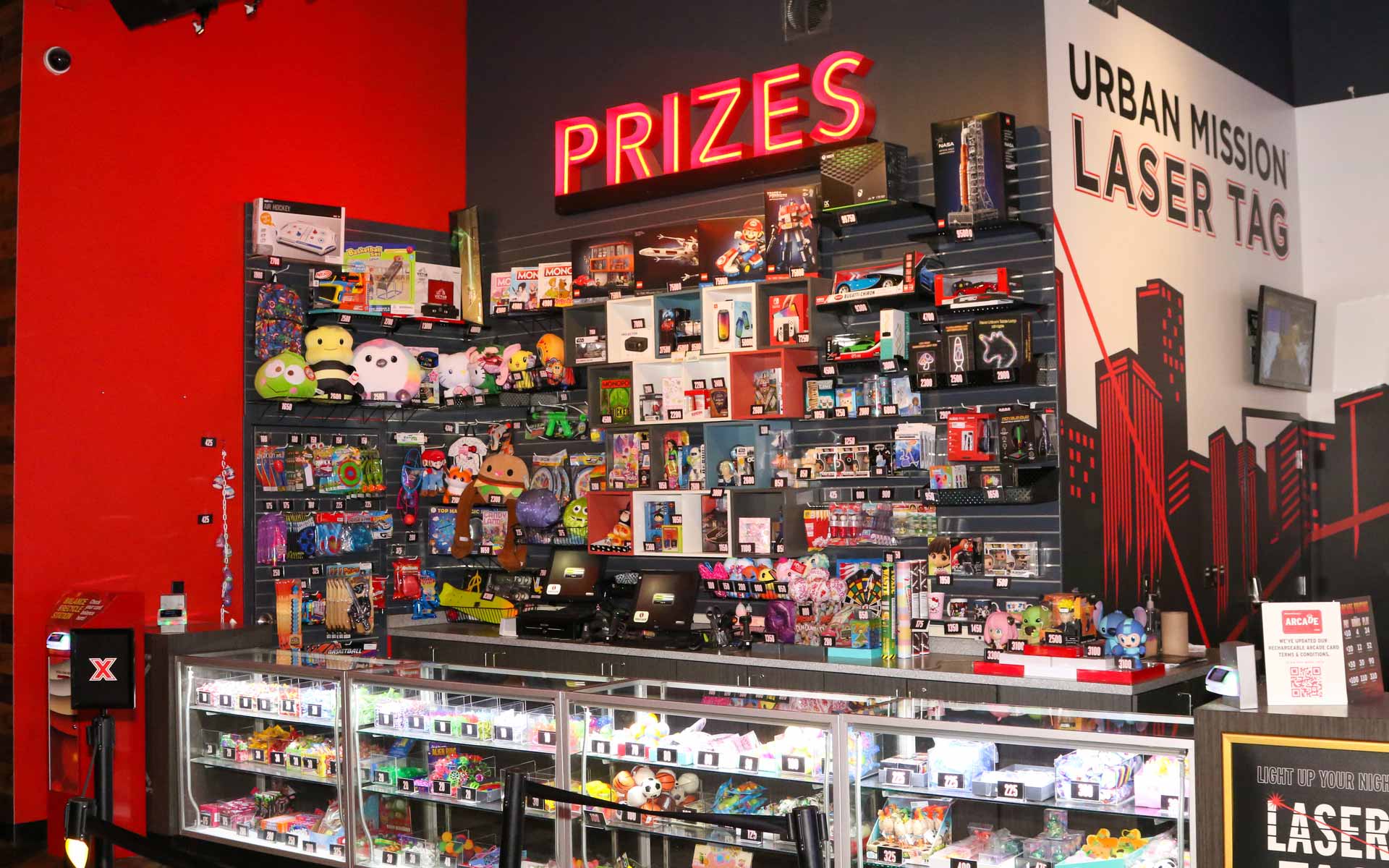 Arcade prize counter with toys, electronics, and display cases under bright “Prizes” signage in a modern entertainment center
