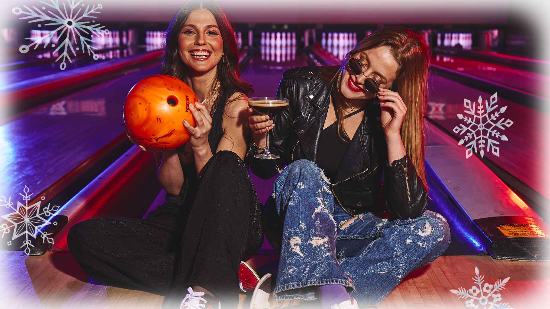 Two women sitting in front of bowling lanes with a bowling ball and a cocktail