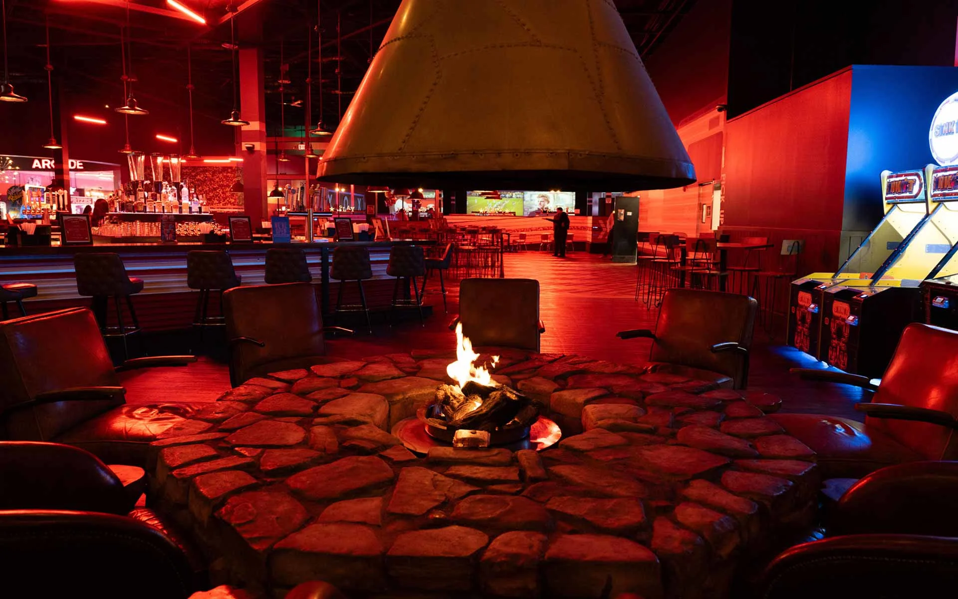 Indoor lounge seating with a circular stone fire pit, surrounded by chairs in a dimly lit Lucky Strike entertainment space.