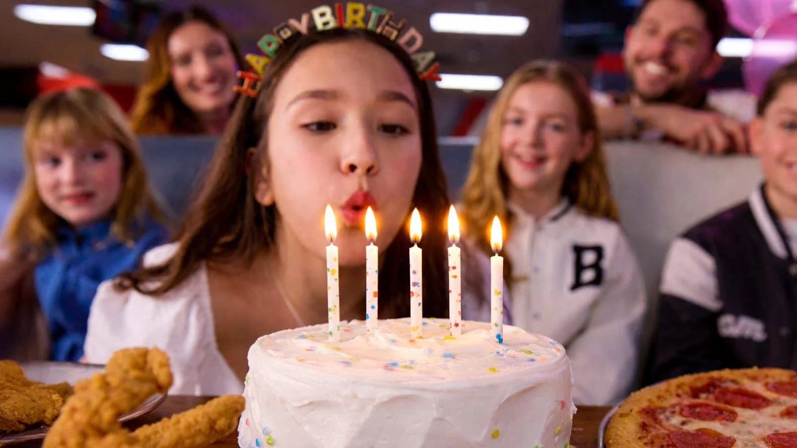 Girl wearing a “Happy Birthday” headband blows out lit candles on a white birthday cake while friends watch at a bowling alley party table with pizza and chicken tenders.