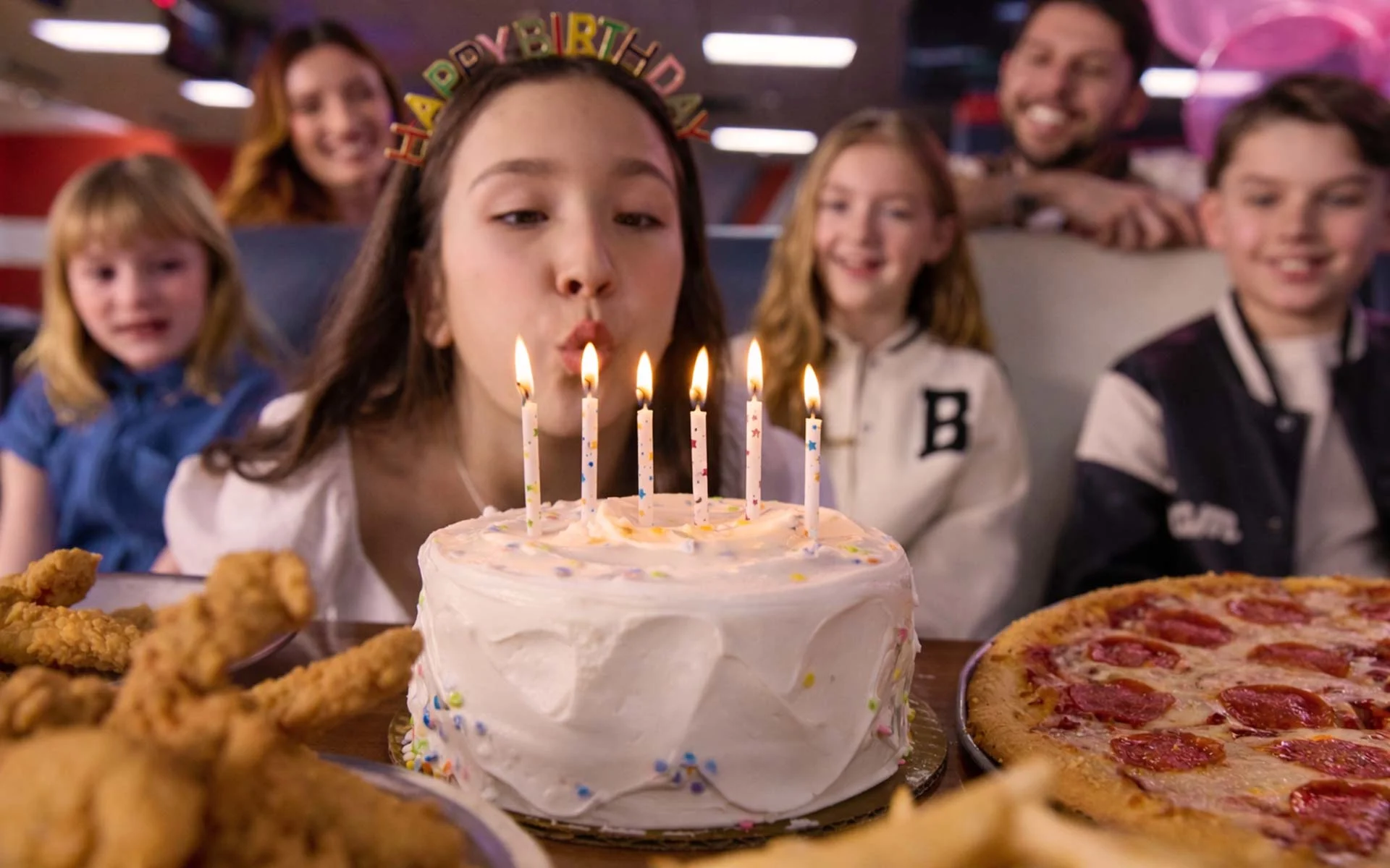 Girl wearing a “Happy Birthday” headband blows out lit candles on a white birthday cake while friends watch at a bowling alley party table with pizza and chicken tenders.
