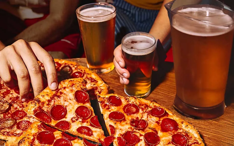 Hand grabbing a slice of pepperoni pizza with a pitcher of beer and two full glasses of beer.