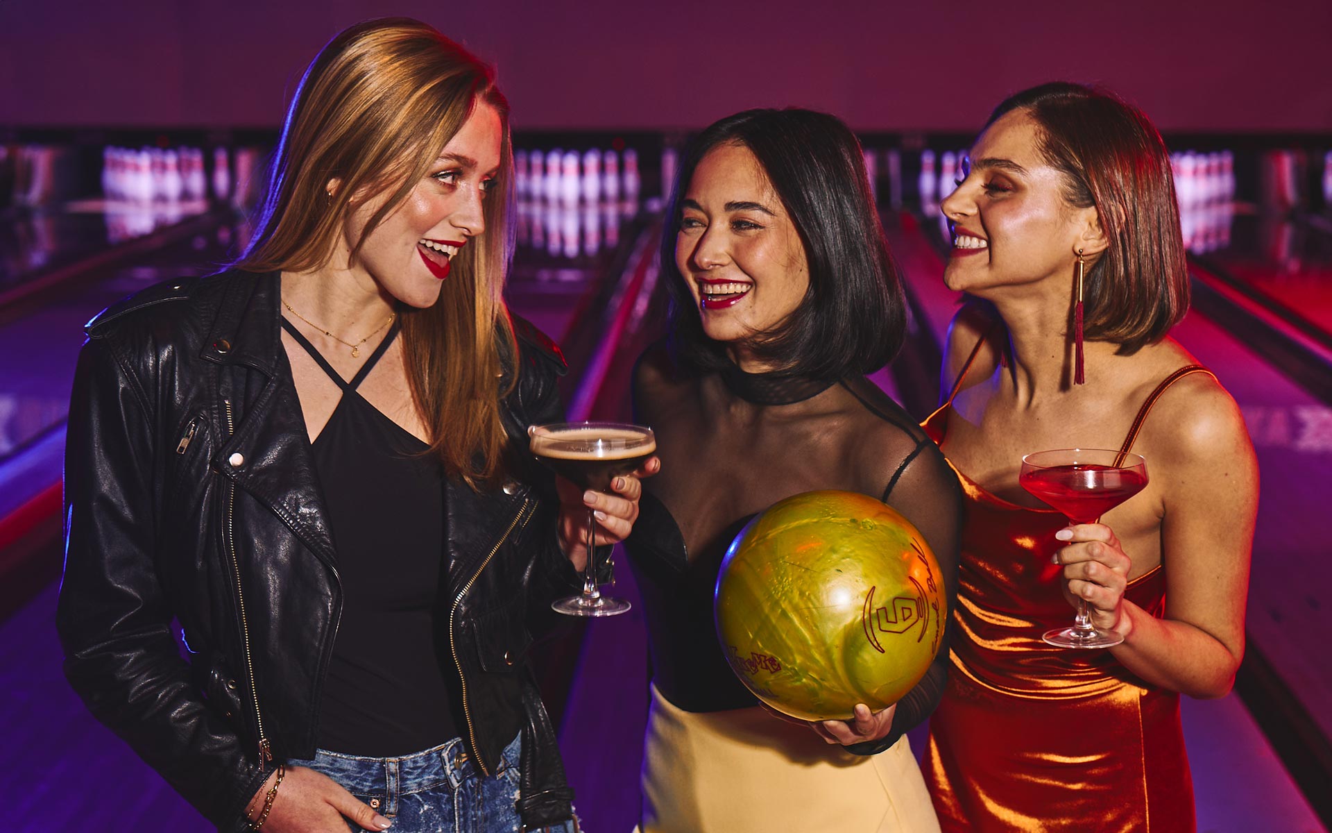 Three girls smiling and holding drinks with bowling lanes in the background.
