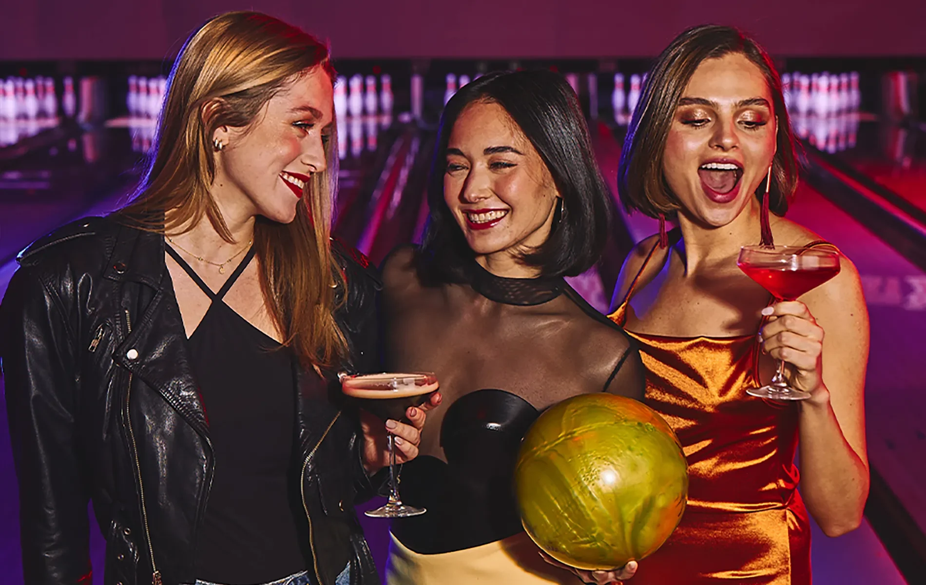 Three girls smiling and holding drinks with bowling lanes in the background.