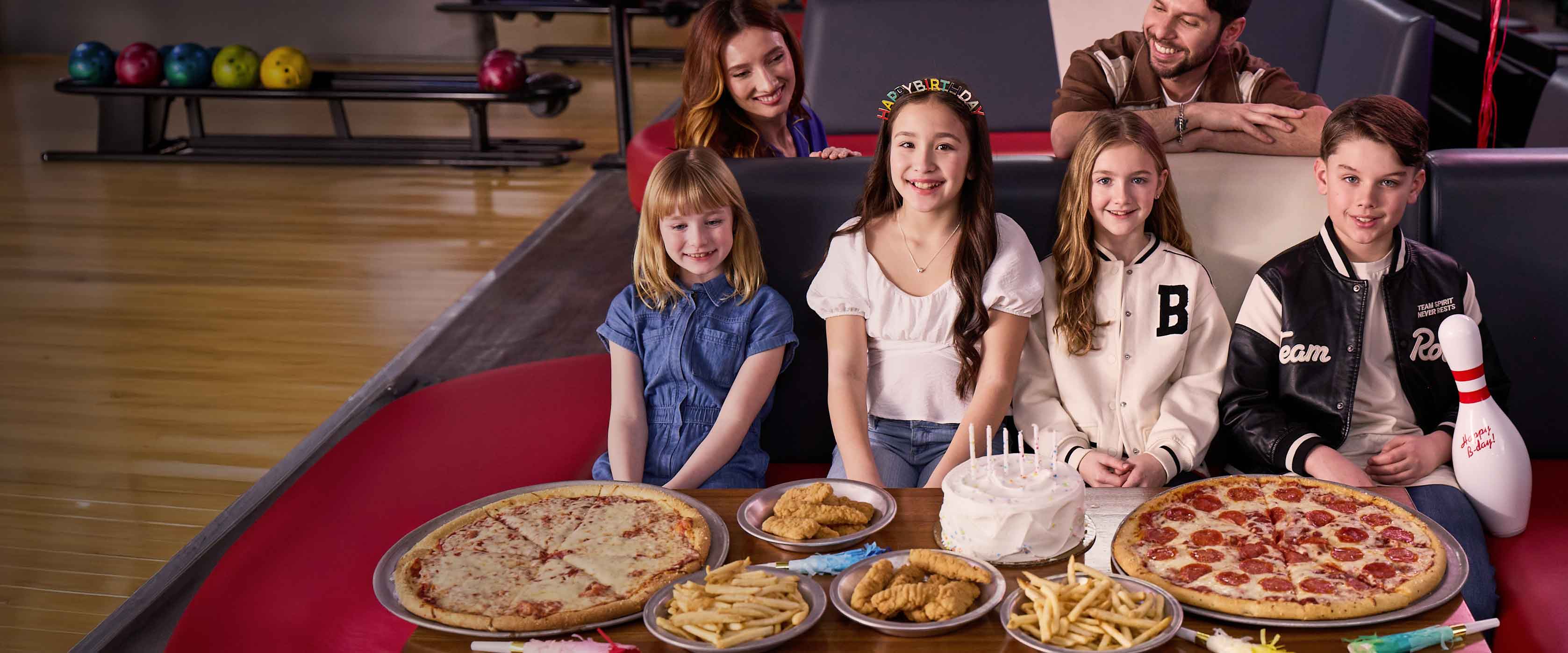 Kids and parents celebrating a birthday party with pizza and cake inside a bowling center.