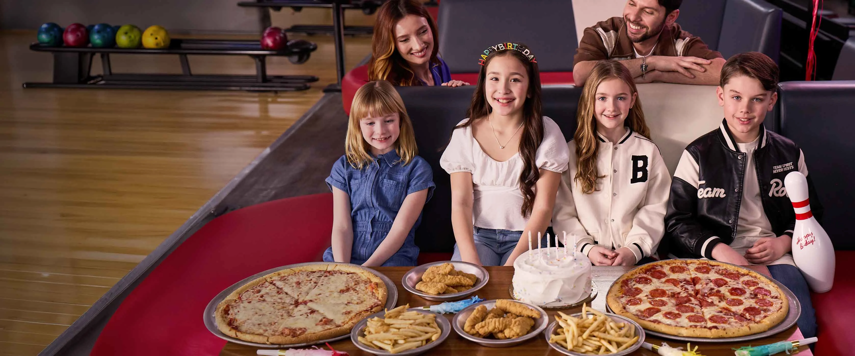 Kids and parents celebrating a birthday party with pizza and cake inside a bowling center.