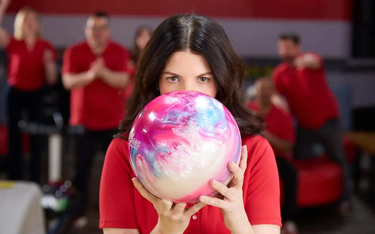 Woman in a red bowling shirt focuses while holding a colorful bowling ball, with teammates cheering in the background at a bowling alley.