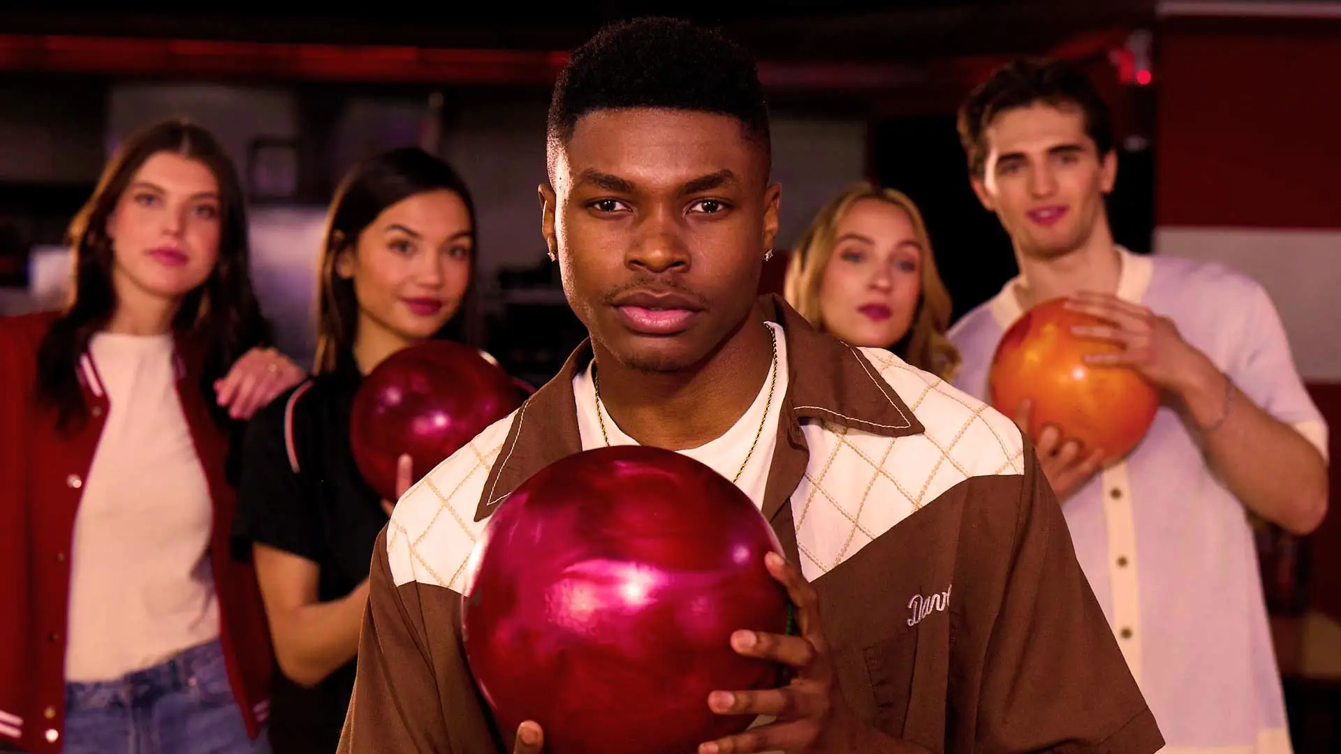Group of young adults at a bowling alley holding colorful bowling balls, with a man in the foreground looking at the camera and friends smiling behind him.