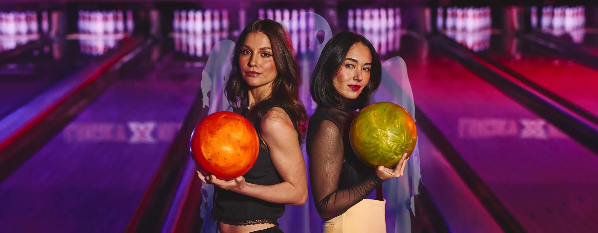 Tow women holding bowling balls with a ghost and bowling lanes in the background