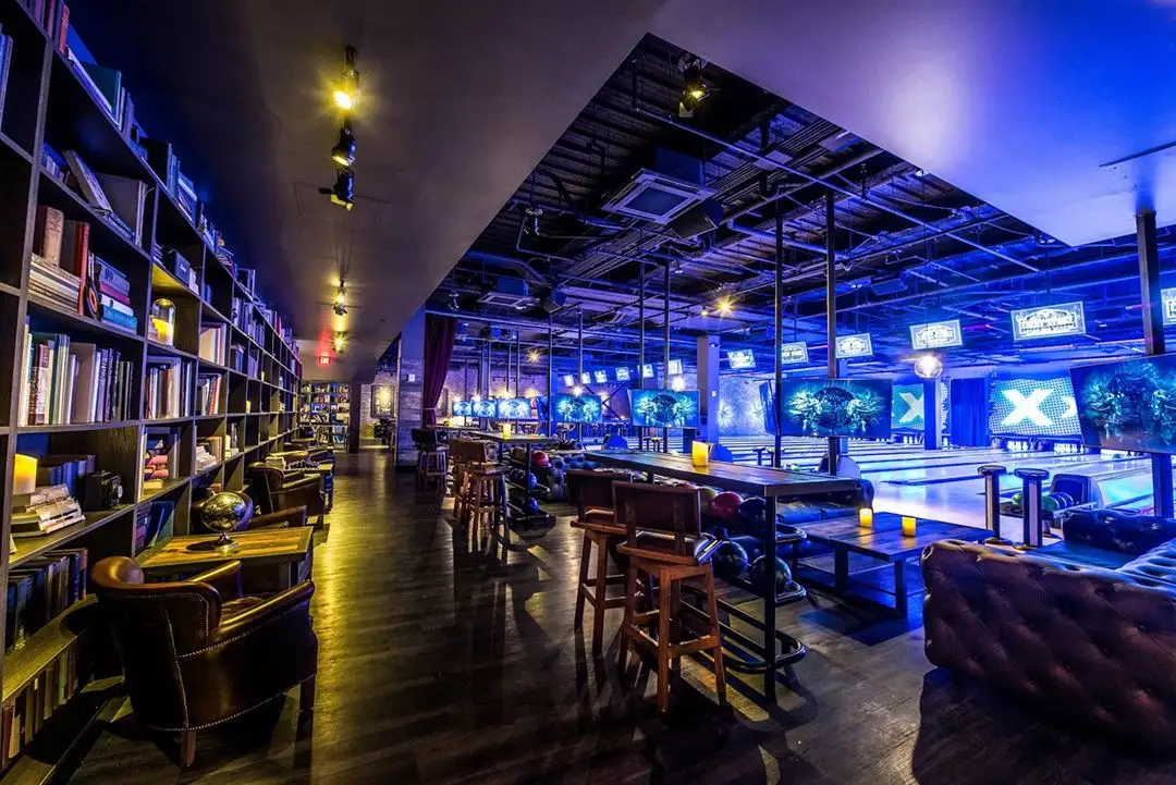 Upscale Lucky Strike bowling lounge with bookshelves, plush seating, high-top tables, and glowing lanes under blue lighting.