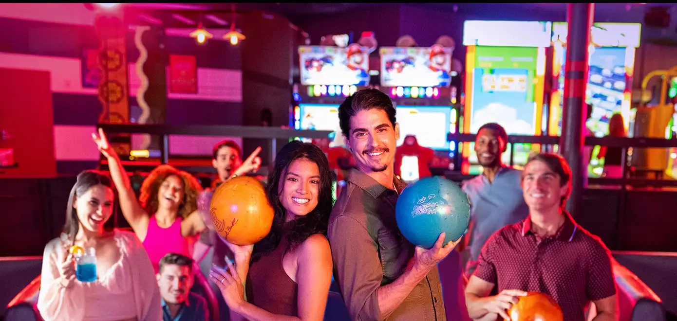 Woman and man holding bowling balls and standing back-to-back in front of a group of other bowlers