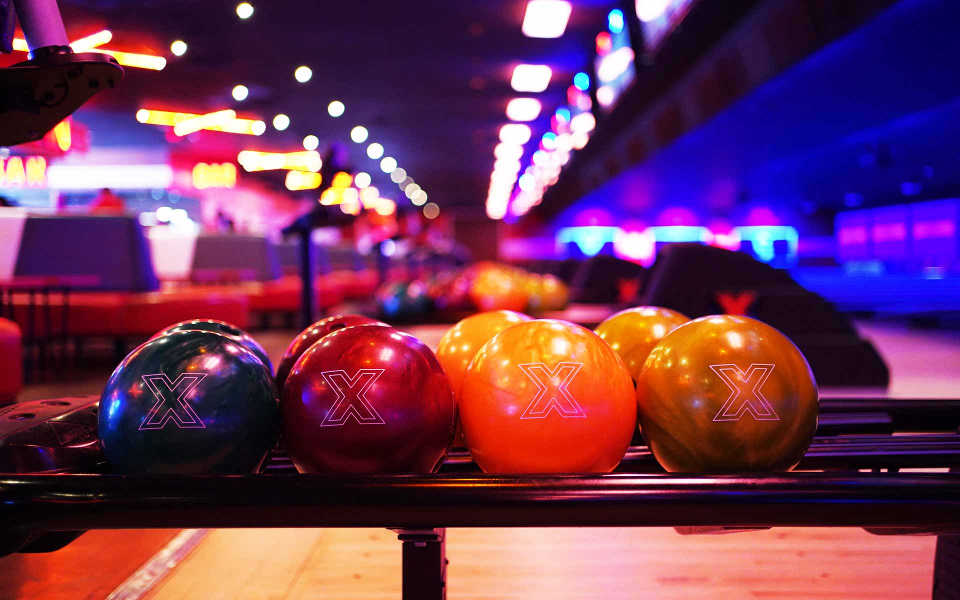 Close-up of bowling balls on a rack with colorful neon lights and lanes softly blurred in the background for a nightlife vibe.