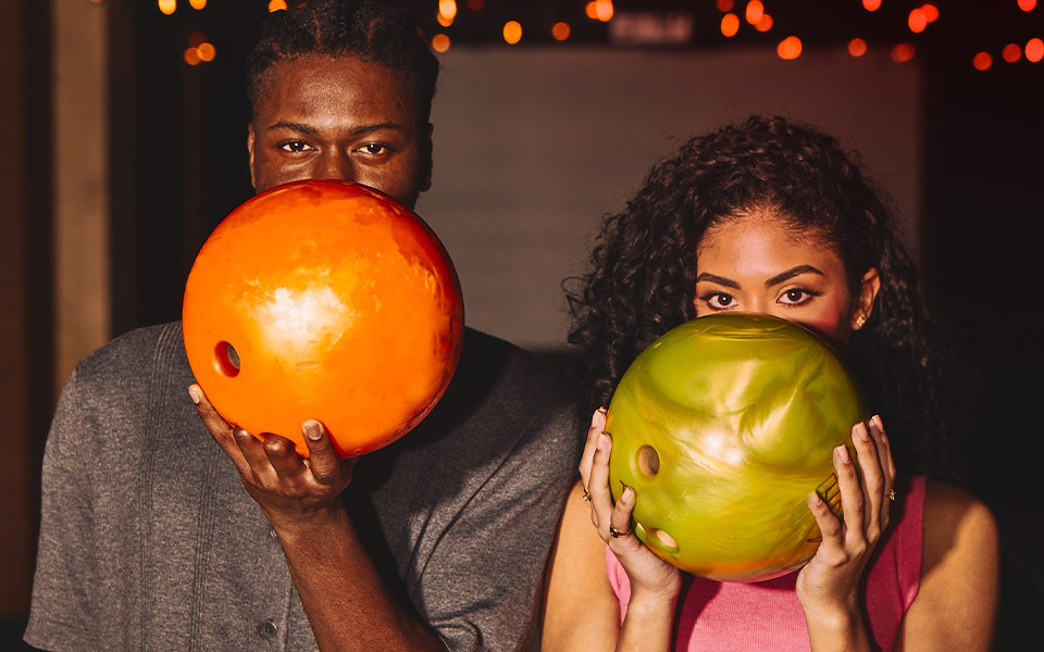 A man and a woman holding up bowling balls coyly. 