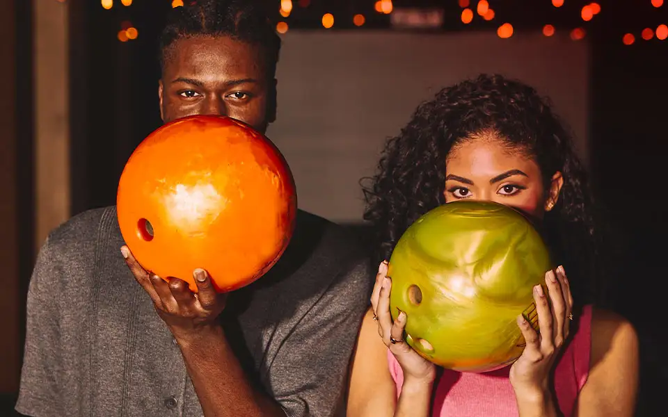 A man and a woman holding up bowling balls coyly.