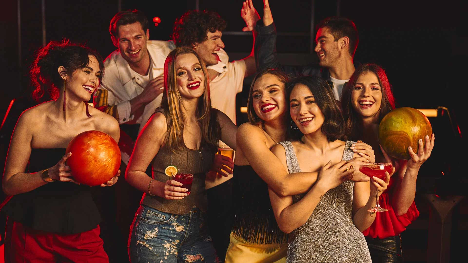 A group of adults smiling and posing with drinks and a bowling ball during a night out at a bowling center.