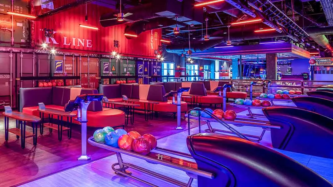 Lucky Strike bowling lanes with red and gray lounge seating, colorful bowling balls, and neon-lit shipping container wall decor