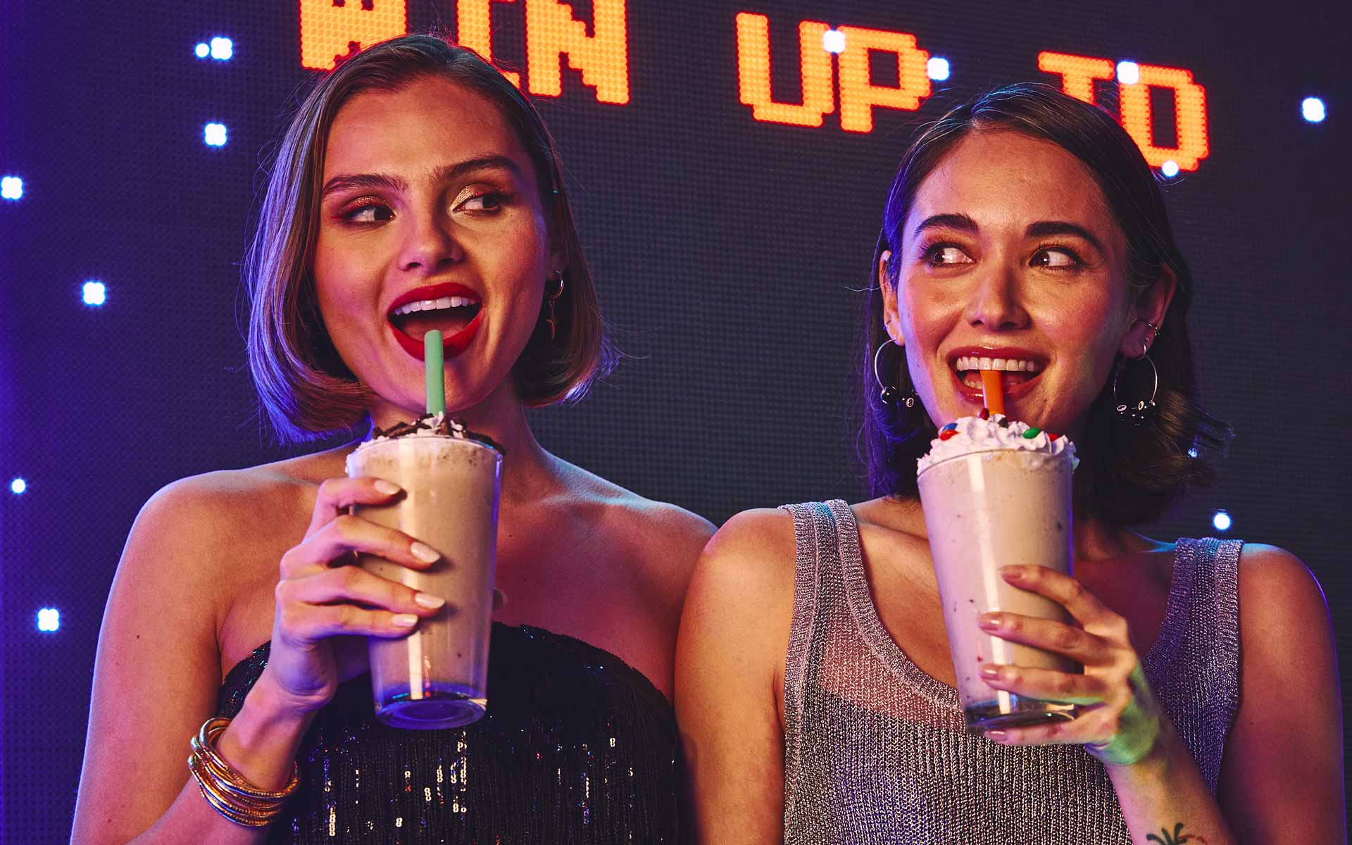 Young women enjoying milkshakes in a neon-lit, upscale bowling lounge with vibrant nightlife atmosphere and colorful lighting