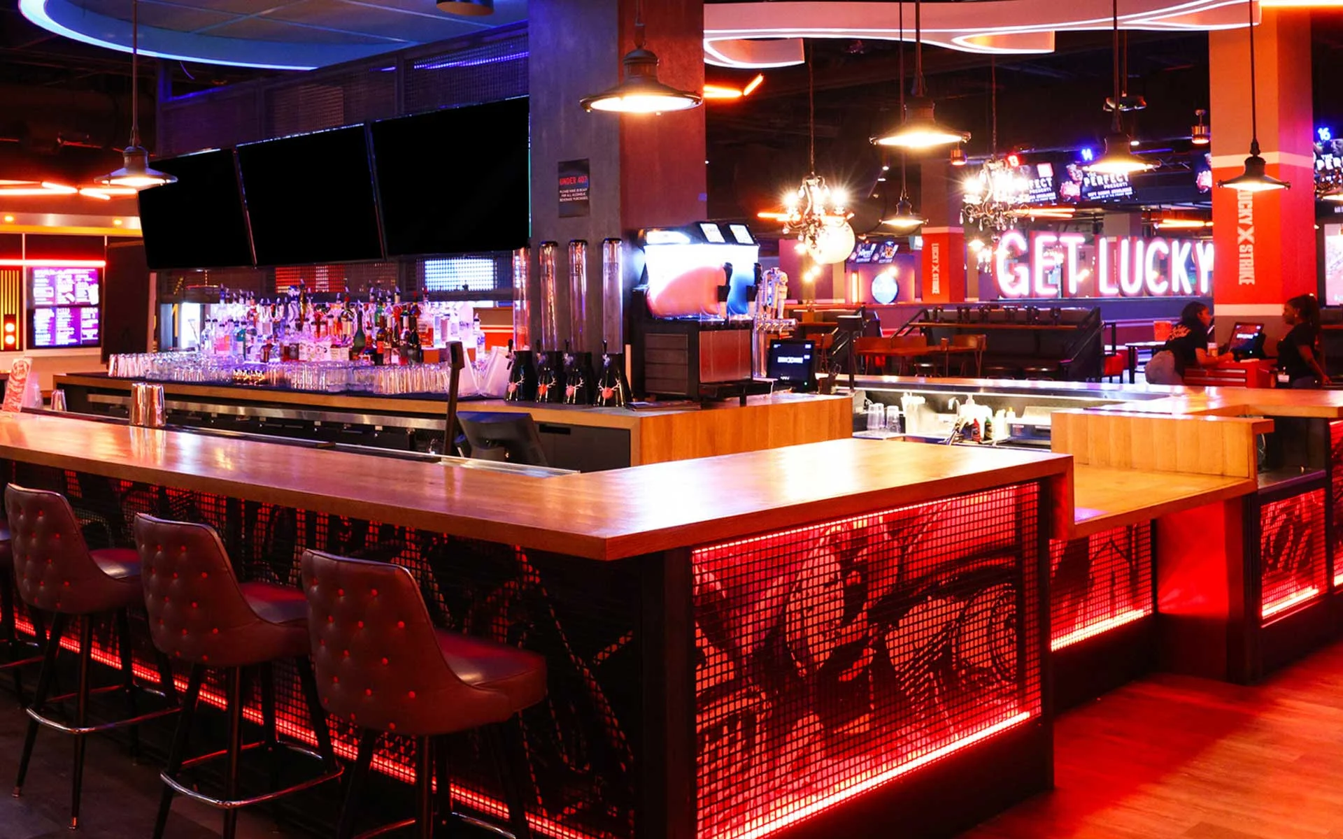Stylish bar area inside a modern bowling venue, featuring a glowing red-lit counter, leather bar stools, multiple TVs, and a fully stocked bar, with bold “Get Lucky” signage and a lively nightlife atmosphere.