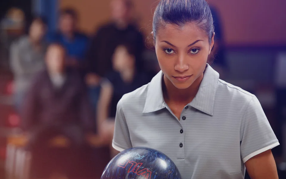 A woman holding a bowling ball in a bowling shirt staring straight at the camera.