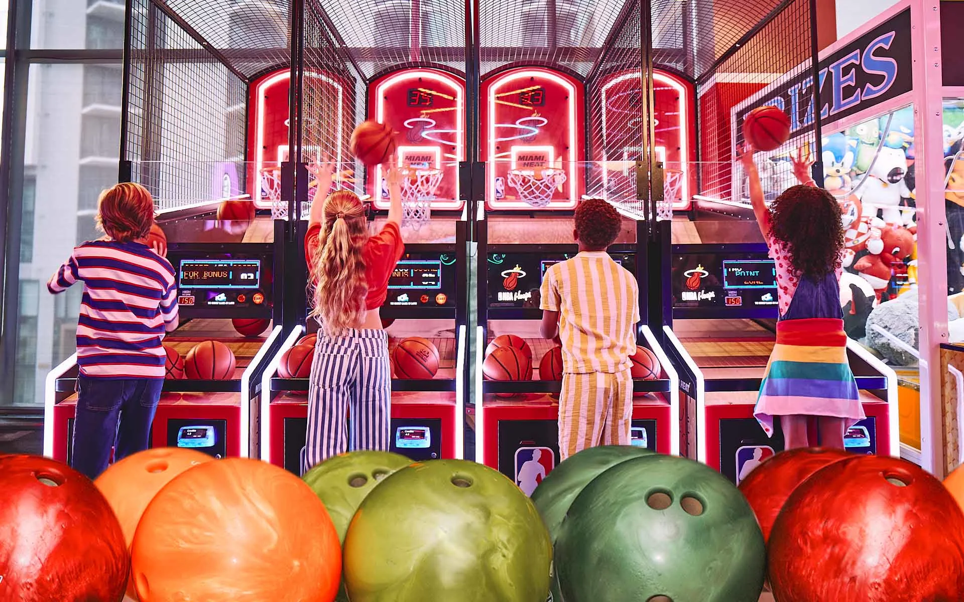 Colorful bowling balls in the foreground with kids playing arcade basketball in the background.