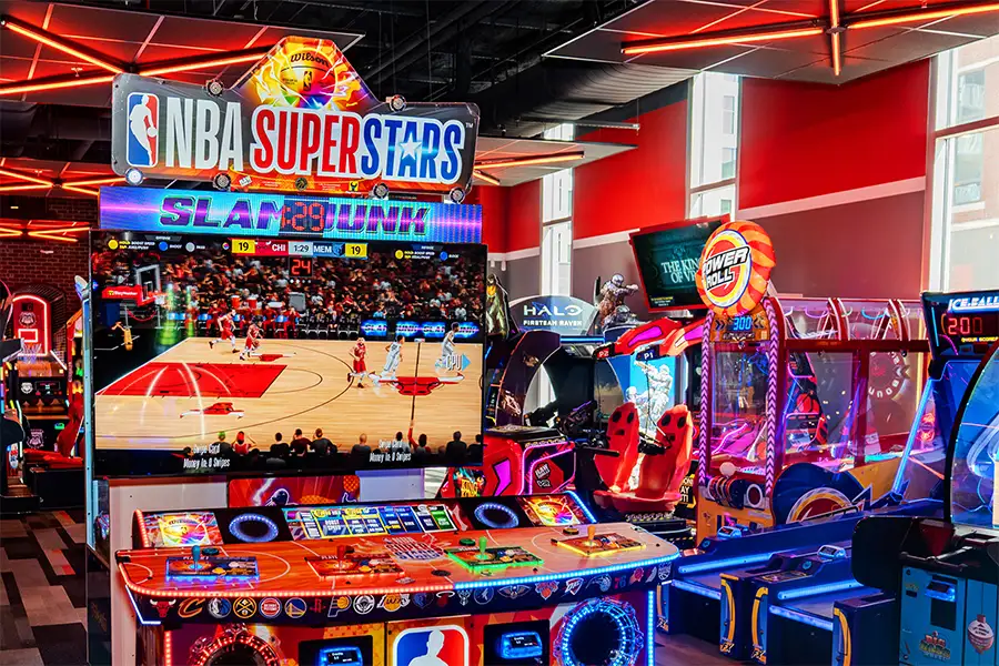 Bright, modern AMF arcade filled with colorful neon lighting and basketball-themed games, featuring a large NBA Superstars Slam Dunk machine surrounded by other interactive arcade attractions and glowing game cabinets.