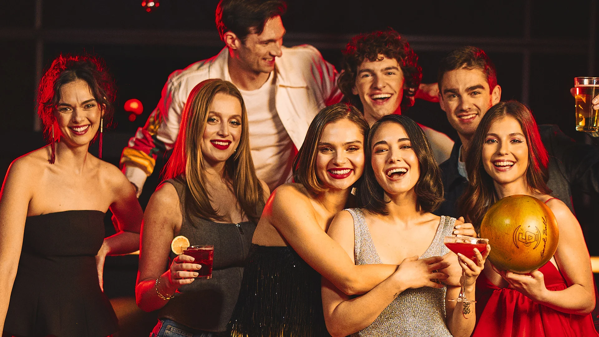 Adults posing together at a bowling night out, holding drinks and a bowling ball in a lively bowling center.