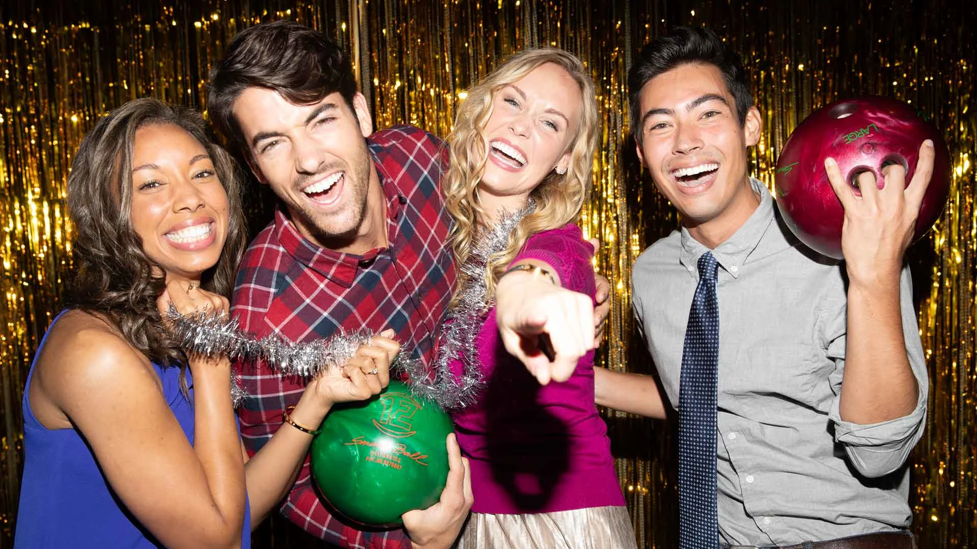 Four smiling adults pose closely together at a festive bowling party, holding bowling balls and wearing dressy outfits in front of a gold glitter backdrop, laughing and pointing toward the camera.
