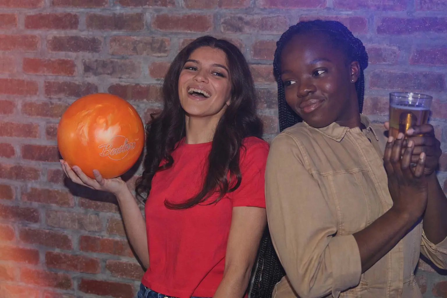 Two women smiling in front of a brick wall with colorful lighting; one holds an orange bowling ball while the other holds a drink.