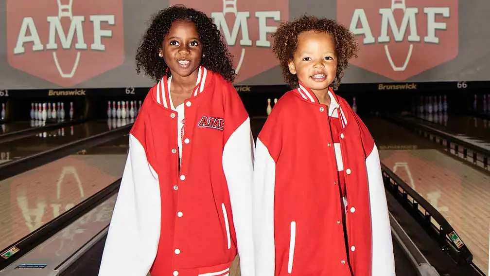 Two kids standing on oversized AMF varsity jerseys in front of a bowling lane at an AMF center.