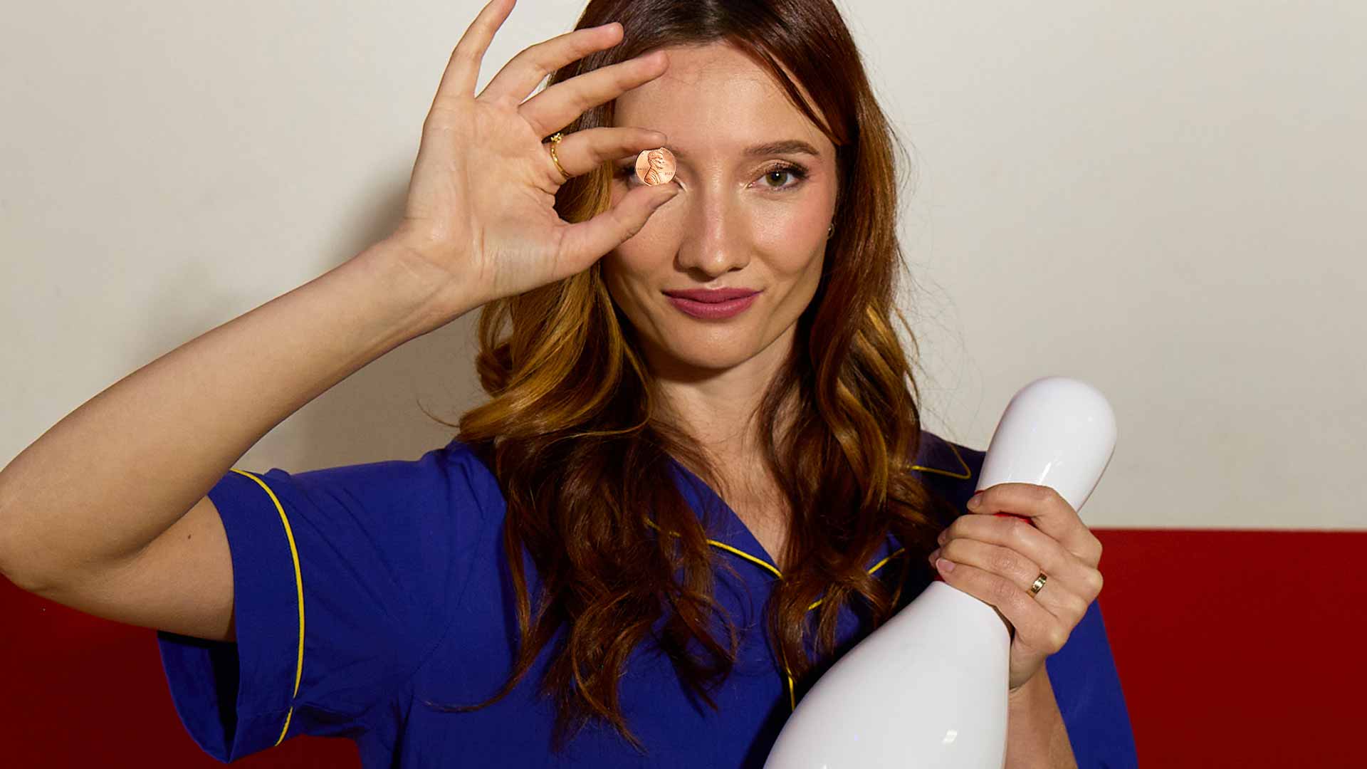 Woman holding a penny in front of her eye and a bowling pin, smiling while posing indoors.