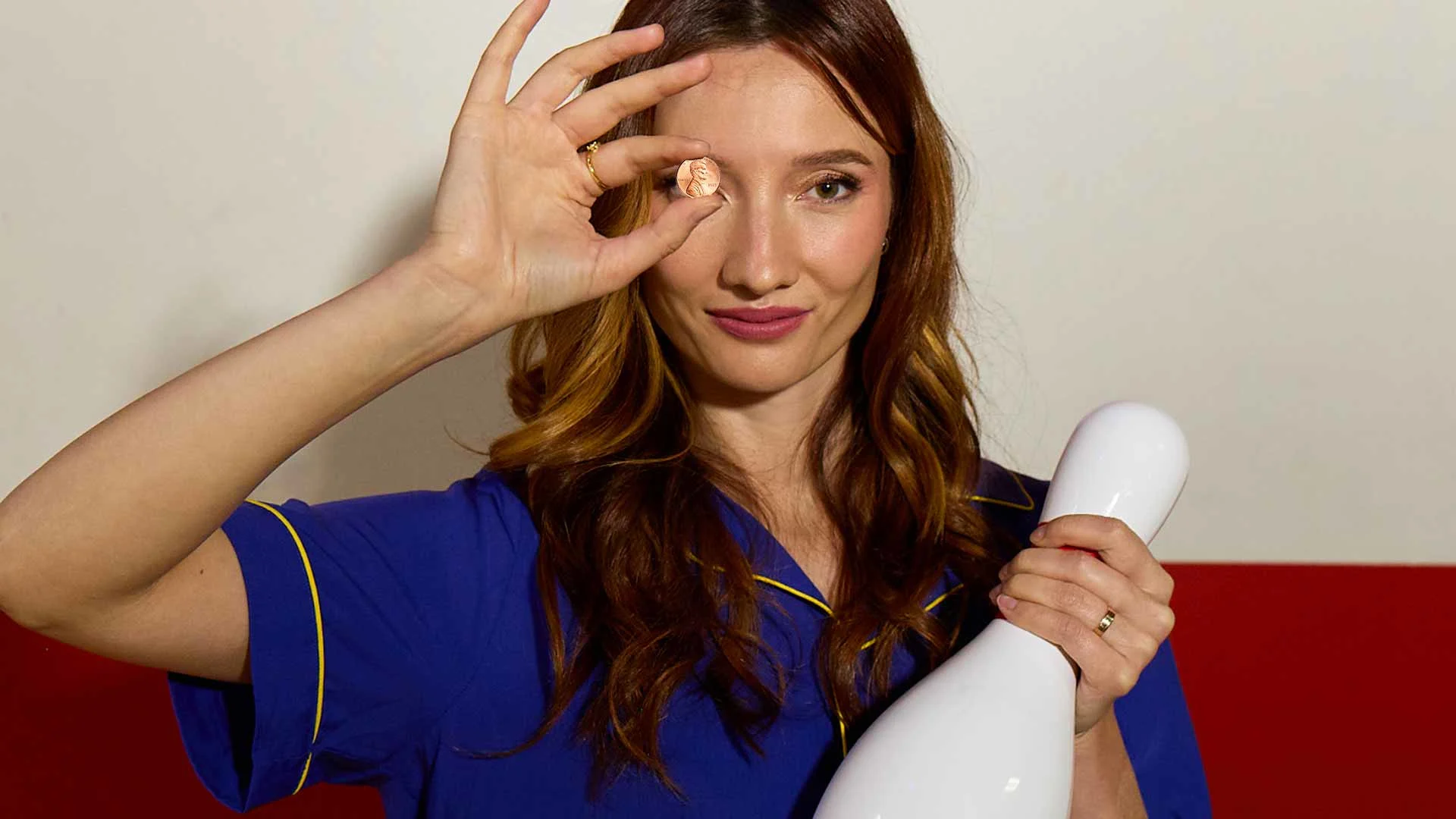 Woman holding a penny in front of her eye and a bowling pin, smiling while posing indoors.