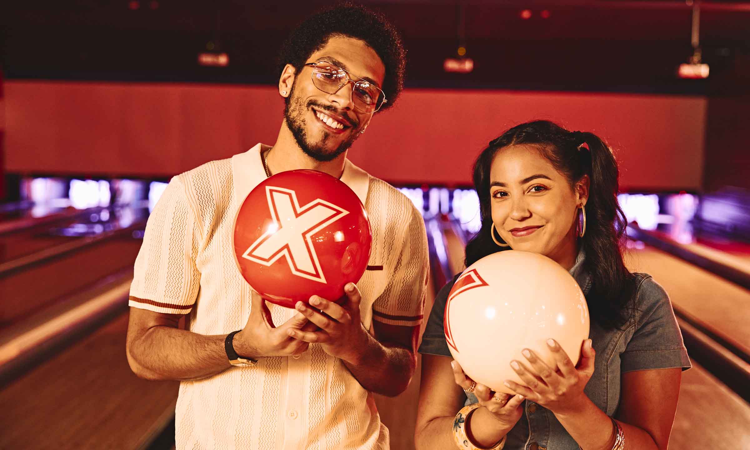 A couple standing on a bowling lane holding bowling balls and smiling toward the camera.