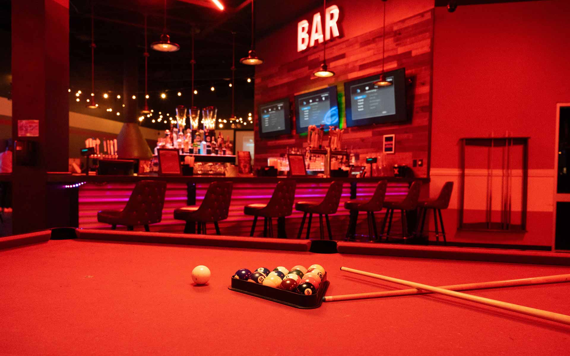 Red-lit billiards table with racked pool balls and cues in front of a stylish Lucky Strike bar with string lights and digital menu screens.
