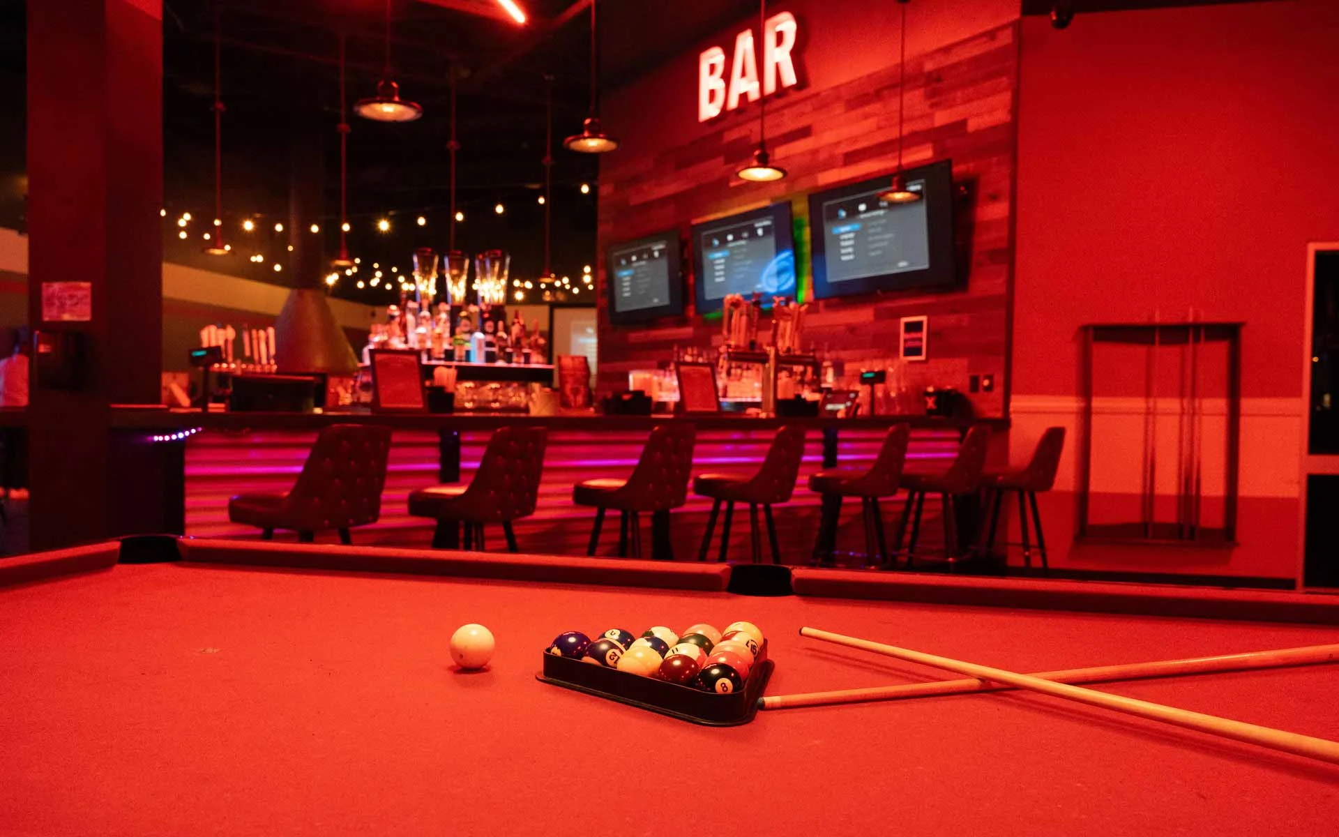 Red-lit billiards table with racked pool balls and cues in front of a stylish Lucky Strike bar with string lights and digital menu screens.