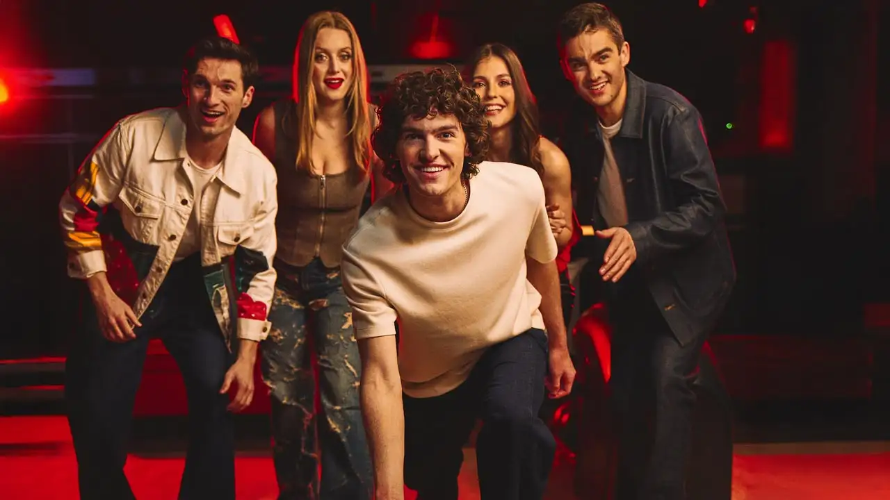 A group of young adults smiling and leaning forward at a bowling alley, with one man in the center about to roll a bowling ball down the lane under red ambient lighting.