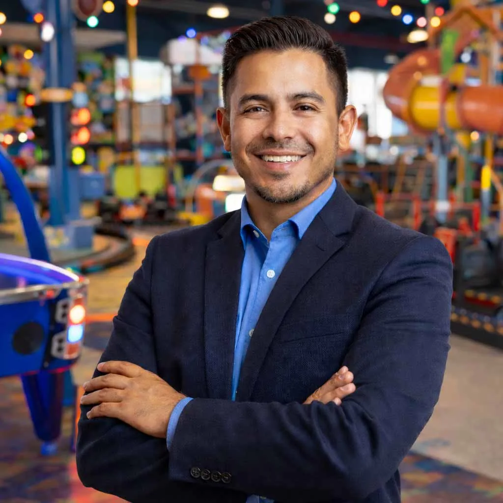 A smiling Latino manager in a blazer and button-down shirt standing with arms crossed inside a family entertainment center with arcade games and attractions in the background.