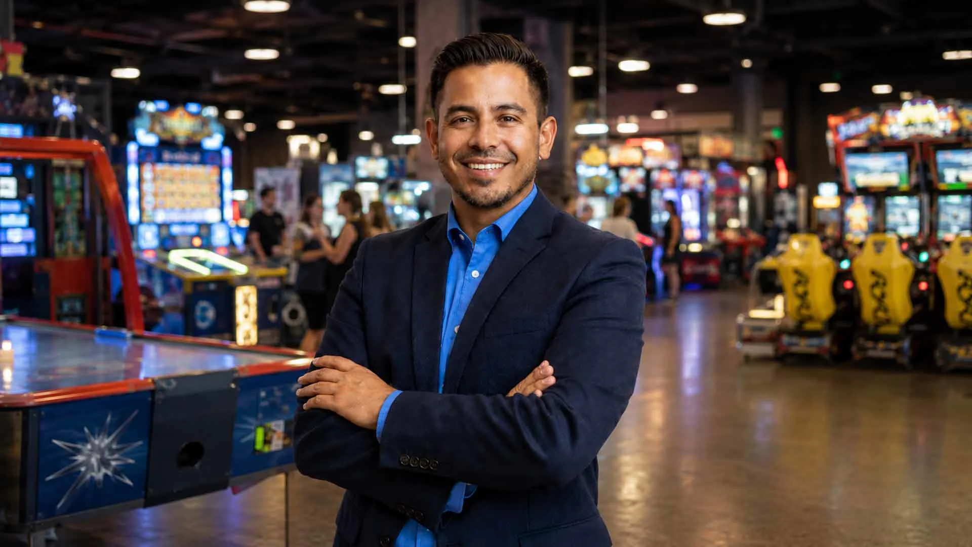 A smiling Latino manager in a blazer and button-down shirt standing with arms crossed inside a family entertainment center with arcade games and attractions in the background.